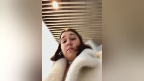 Media: Video of a woman with light skin and brown hair, wearing a white fur coat, standing in a modern room with a wooden ceiling.