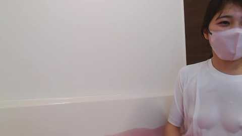 Media: Video of a young Asian woman with short black hair, wearing a white T-shirt and face mask, partially submerged in a pink bathtub against a plain white wall.