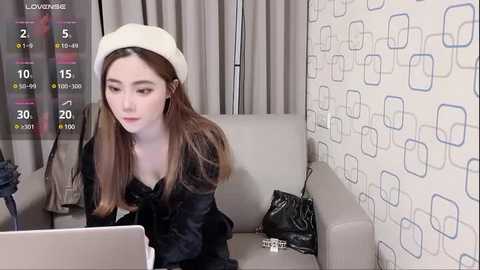 Media: Video of a young Asian woman with long brown hair, wearing a white beret and black blouse, seated in a modern living room with geometric-patterned wallpaper, using a laptop.