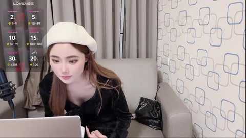 Media: Video of an Asian woman in a beige beret and black coat, seated on a beige couch, using a laptop in a room with a patterned wallpaper.