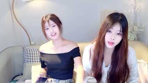 Media: Video of two young Asian women with straight, long brown hair, wearing casual off-shoulder black tops, seated on a beige couch in a softly lit room with floral decorations.