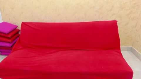 Media: Video of a bright red sofa covered with a matching red fabric, positioned against a beige textured wall, with a stack of pink and purple cushions on the left.