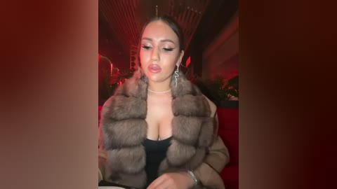 Media: Video of a fair-skinned woman with light brown hair, wearing a fur-lined coat, black top, and silver earrings, sitting in a dimly lit bar with red lighting.