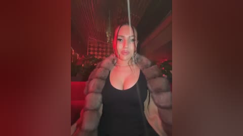 Media: Video of a confident, light-skinned woman with medium-length blonde hair, wearing a black dress and fur coat, against a dimly lit, red-lit background with a high-end interior setting.