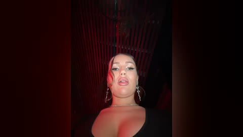 Media: Video of a light-skinned woman with medium-length hair, wearing large hoop earrings and a low-cut black top, in a dimly lit room with red lighting, emphasizing her makeup and sultry expression.