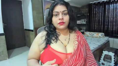 Media: Video of a plus-sized South Asian woman with long black hair, wearing a red sari and a sheer red bra, sitting on a chair in a cluttered bedroom with a bed and bookshelf.