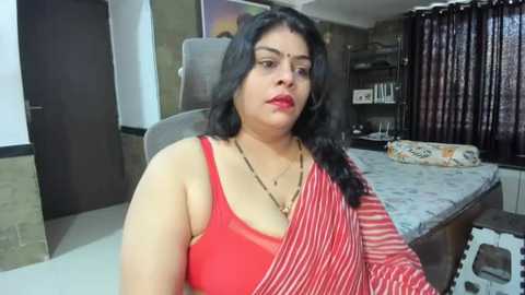 Media: Video of a middle-aged South Asian woman with dark hair, wearing a red and white striped sari, seated in a modern bedroom with a large bed, dark curtains, and a colorful throw pillow.