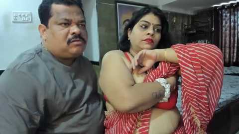 Media: Video of a South Asian man in a grey shirt and a woman in a red and white striped sari, both looking serious in a dimly lit room with a bed and curtains in the background.