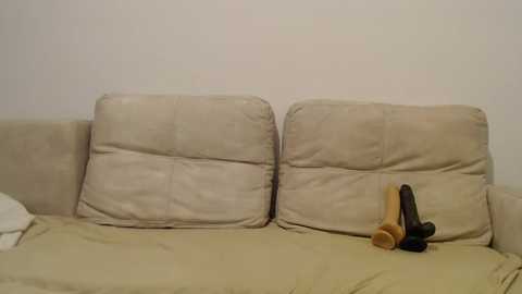 Media: Video of a beige couch with two beige cushions, featuring a tan dildo and a black vibrator on the cushion on the right. The background is a plain white wall.