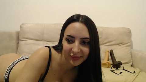 Media: Video of a young woman with long black hair, wearing black lingerie, lying on a beige couch, holding a dildo, in a casual indoor setting.