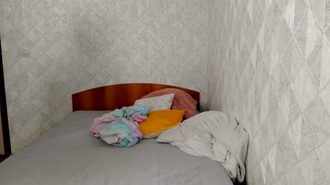 Media: Video of a messy, small bedroom with a single bed covered in a colorful, patterned blanket. The walls are covered in white, textured wallpaper. The room appears neglected and cluttered.