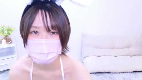 Media: A video of an Asian woman with short brown hair, wearing a pink face mask and bunny ears, seated in a minimalist, white-walled room with a plush beige armchair and a small table with a potted plant.