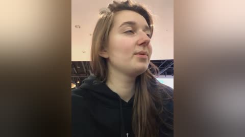 Media: Video of a young woman with light skin and long, light brown hair, wearing a black hoodie, gazing upwards with a contemplative expression, indoors with a blurred background.