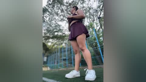 Media: Video of a young woman in a maroon sports outfit, white sneakers, and a ponytail, stretching outdoors on a grassy field behind a blue fence.