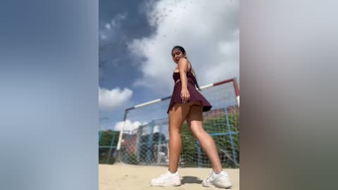 Media: Video of a young woman with long dark hair, wearing a short, dark purple dress, standing on a basketball court with a chain-link fence in the background under a partly cloudy sky.