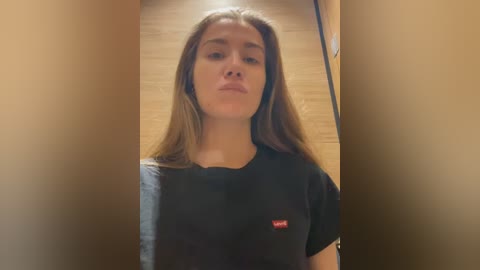 A video of a young woman with light skin, straight brown hair, and a casual black t-shirt featuring a small red logo, standing in a narrow, dimly lit hallway with wooden walls.