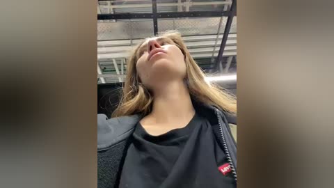 Video of a young woman with long, straight blonde hair, wearing a black jacket over a black shirt, standing indoors with a metallic, industrial ceiling and blurred background.