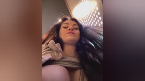 Media: Video of a young woman with long dark hair, wearing a grey sweater, lying on a bed, eyes closed, in a dimly lit room with a hanging chandelier.