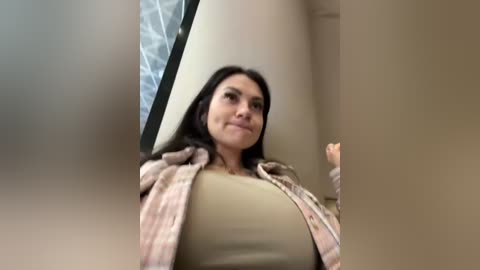 Media: Video of a woman with shoulder-length dark hair, wearing a beige top and plaid shirt, standing in a beige-walled room with a large window.