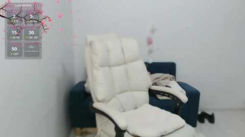 Media: Video of a cream leather recliner chair with a dark blue cushion, placed in a white room with a digital thermometer display showing 27\u00b0C. A beige blanket is draped over the chair, and a white wall with pink cherry blossom decals is visible.