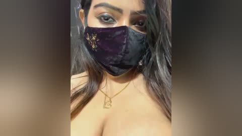 Media: Video of a South Asian woman with medium skin tone, long dark hair, wearing a black face mask, purple velvet mask with gold embroidery, and a gold necklace with a rectangular pendant.