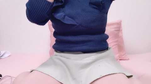 Media: Video of a person, possibly female, wearing a blue sweater and light-colored shorts, sitting on a pink bed with a pillow, with a white object in the background.