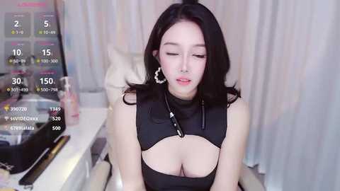 Media: Video of an East Asian woman with long black hair, wearing a revealing black crop top with cutouts, sitting in a modern room.