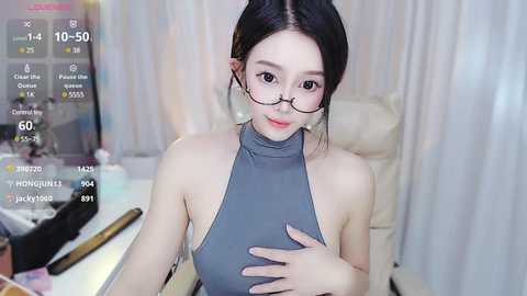 Media: A video of an East Asian woman with fair skin, wearing glasses, a gray halter top, and black hair in a bun. She's seated at a desk, with a virtual camera overlay showing her age, location, and stats.