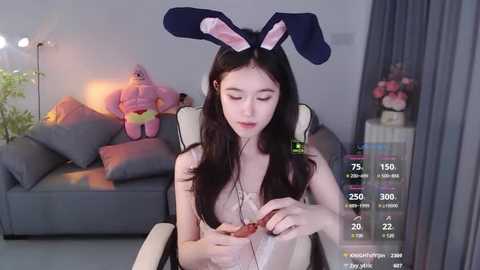 Media: Video of an Asian woman in a gray chair, wearing bunny ears and a white top, eating strawberries, with a plush pink rabbit and gray sofa in the background.