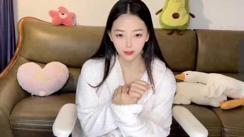 Media: Video of an Asian woman with long black hair, wearing a white robe, sitting on a brown leather couch. Background includes plush toys: a pink bear, a yellow duck, and a white swan.