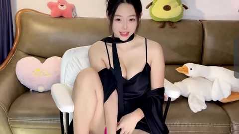 Media: Video of an Asian woman with black hair in a bun, wearing a black satin dress, sitting on a beige couch, surrounded by plush toys including a green frog, a white rabbit, and a pink heart.