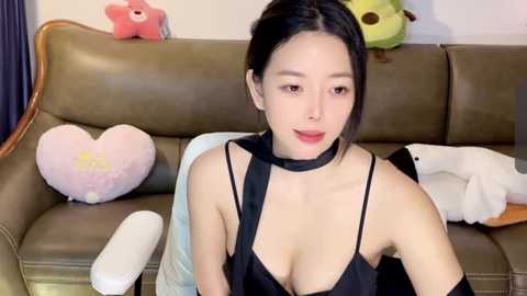 Media: Video of an Asian woman with long black hair, wearing a black choker and revealing black top, sitting on a beige leather sofa. Background includes plush toys, heart-shaped pillows, and a white stuffed animal.