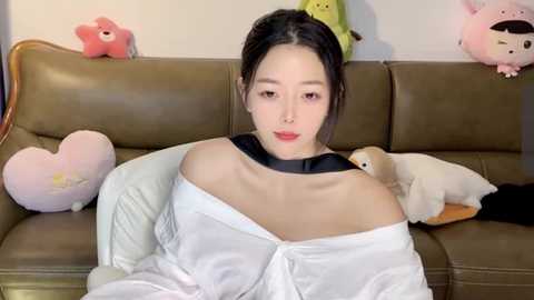 Media: Video of a young East Asian woman with fair skin and dark hair, wearing an off-shoulder white blouse, seated on a brown leather couch. Surrounding her are plush toys, including a pink heart, a green dragon, and a beige stuffed animal.