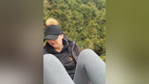 Media: Video of a blonde woman with a ponytail, wearing a black baseball cap, dark jacket, and light gray leggings, sitting outdoors against a backdrop of dense green foliage.