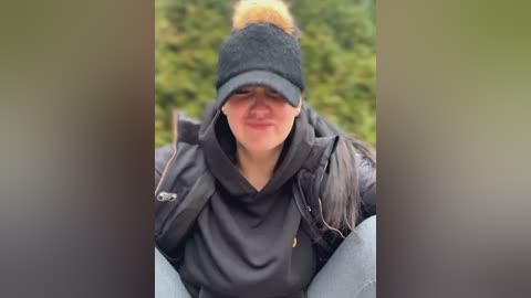 Media: Video of a Caucasian woman with light skin and blonde hair, wearing a black beanie, dark jacket, and jeans, sitting outdoors against a blurred green background.