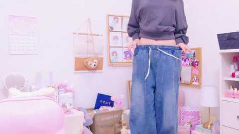 Video of a person pulling up baggy blue jeans in a pastel-themed, cluttered bedroom with pink and white decor, including a stuffed bear, calendar, and shelves.