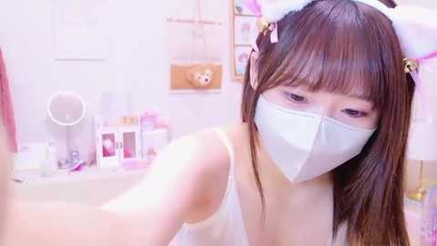 Media: Video of a young East Asian woman with straight, shoulder-length brown hair and light skin, wearing a white face mask, white tank top, and pink cat ears. Background features a bathroom with toiletries and a small teddy bear.