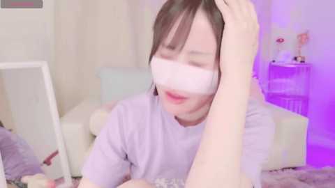 Media: A video of a young Asian woman with light skin, wearing a white mask, smiling, touching her hair, in a pastel-colored room with a bed and a shelf with toys.