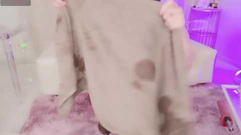 Media: A blurred video of a person holding a large, tattered, light-colored garment with dark stains, in a dimly lit room with purple lighting and a fluffy pink rug.