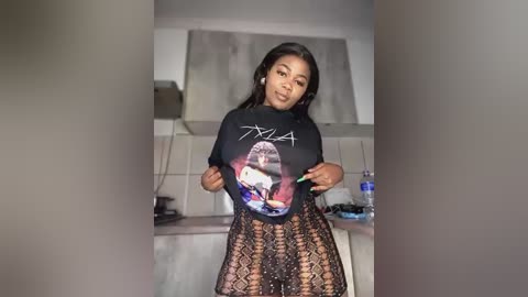 Media: Video of a Black woman with medium skin tone and long straight hair, wearing a sheer black top with a graphic print, and a black lace skirt. She stands in a modern kitchen with white cabinets and a blue water bottle.