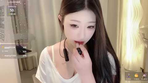 Media: A video of an East Asian woman with long black hair, light skin, and a slender build, wearing a white off-shoulder top, applying makeup in a dimly lit room.