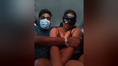 Media: Video of a South Asian woman with medium skin tone and dark hair, wearing a surgical mask and revealing black lingerie, seated on a gray couch, with a man in a green shirt behind her.