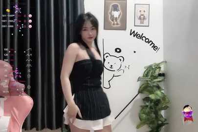 Media: Video of an Asian woman with long black hair, wearing a black strapless top and white shorts, in a cozy room with a teddy bear mural, plants, and black curtains.