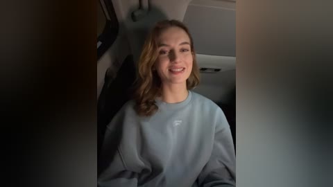 Media: Video of a young woman with light skin and wavy, shoulder-length blonde hair, smiling and wearing a light gray hoodie. She's seated in the backseat of a car, with dim, muted lighting.