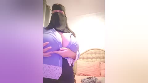 Media: A video captures a plus-sized woman with a black hijab, large breasts, and a black top, holding her buttocks in a bedroom with a bed and wall-mounted lamp.