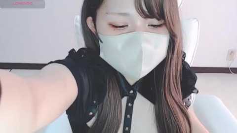 Media: Video of an East Asian woman with long, straight brown hair, wearing a white face mask and black blouse, seated in a dentist's chair with white walls and a wall-mounted device in the background.