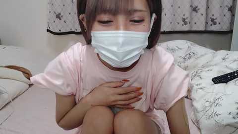 Media: Video of an Asian woman in a pink shirt, wearing a face mask, with her hands on her chest, sitting on a bed with white floral sheets and a black remote control.