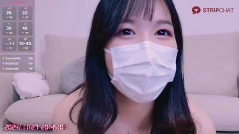 Media: A young Asian woman with long black hair, wearing a white surgical mask, sits on a beige couch in a living room, with a smartphone screen displaying \"STRICTCHAT\" and a thermometer reading.