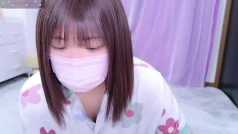 Media: Video of an Asian woman with straight brown hair and bangs, wearing a pink face mask and a floral-patterned white kimono, kneeling on a light gray floor, indoors with pastel-colored curtains and a white dresser in the background.