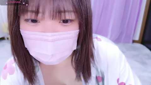 Media: Video of an Asian woman with straight brown hair, wearing a white face mask and a floral-patterned robe, sitting on a light gray carpet in a pastel-colored room.